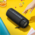 A black portable Bluetooth speaker with a textured fabric cover and a top control panel, shown on a bright yellow surface with a hand reaching toward it. A glass with a lime, a lemon slice, and small colorful pieces are placed around it as styled props.
