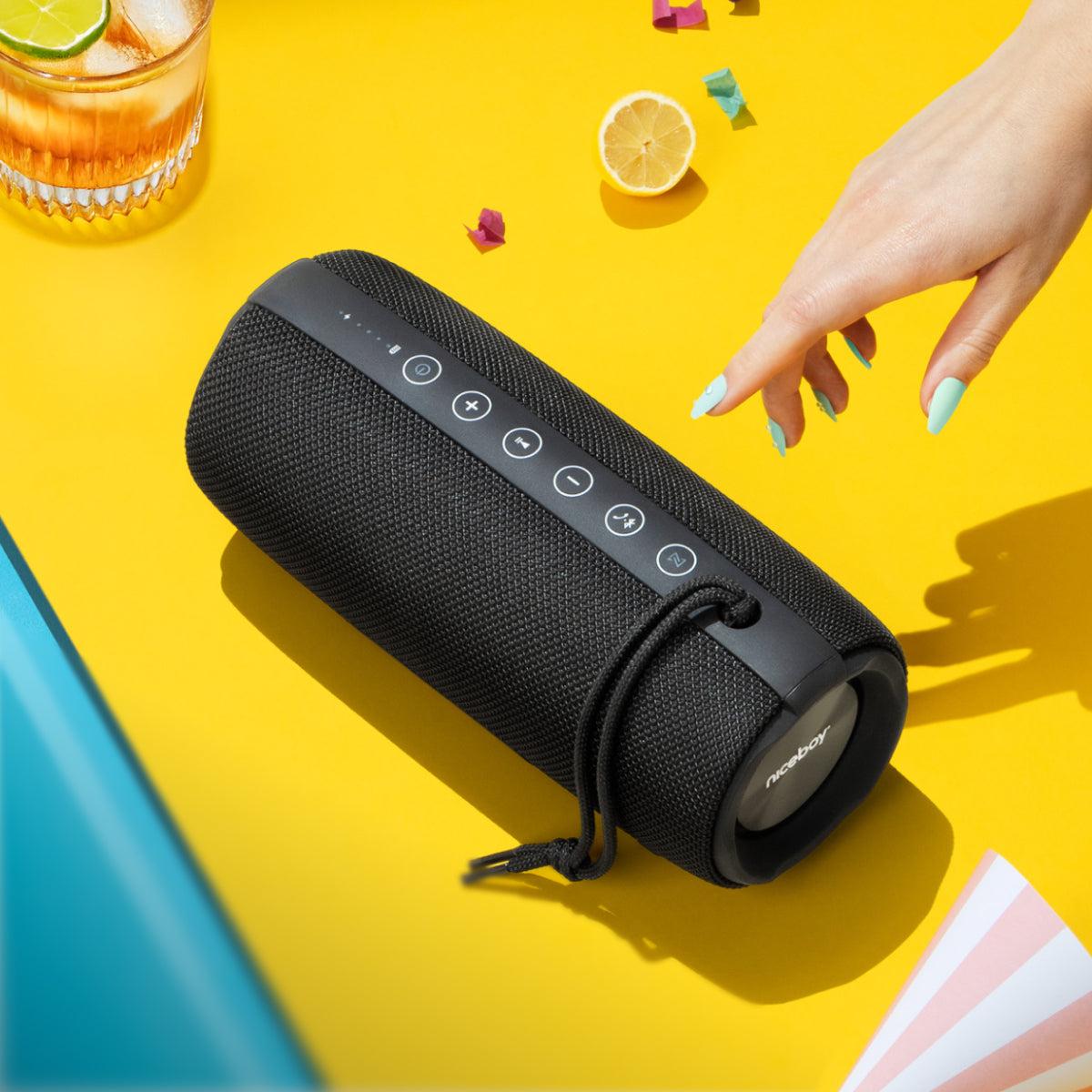 A black portable Bluetooth speaker with a textured fabric cover and a top control panel, shown on a bright yellow surface with a hand reaching toward it. A glass with a lime, a lemon slice, and small colorful pieces are placed around it as styled props.